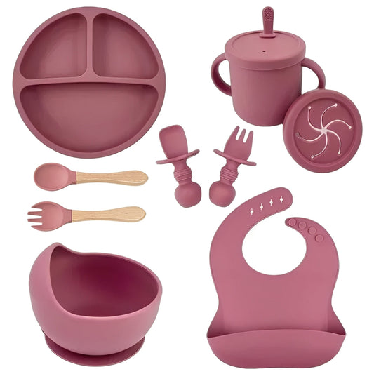 Pink silicone baby feeding set including plate, cup, bowl, utensils, and bib on a white background